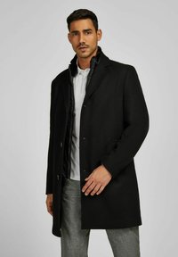 Cappotto nero con colletto a revers e chiusura a bottoni, dotato di una fodera interna trapuntata, tessuto in lana morbido e tasche laterali.