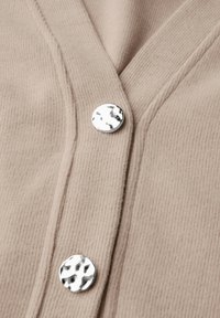 Beige ribbad kofta med V-ringning, prydd med silverfärgade, texturerade knappar. Tyget har ett mjukt, varmt utseende och lätt stretch.
