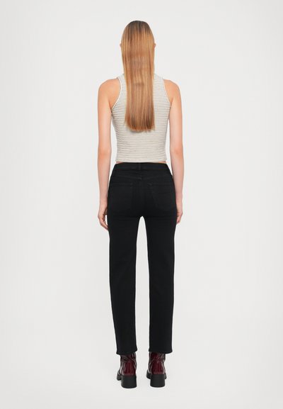 Jeans in denim nero con taglio dritto, indossati con una canotta a righe. I capelli del modello sono lunghi e lisci, abbinati a stivali d'ankle lucidi color borgogna.