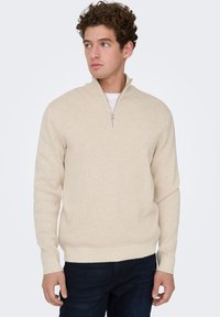 Beige quarter-zip sweater in soft knit fabric, featuring a tapered collar and ribbed cuffs, paired with dark denim pants.