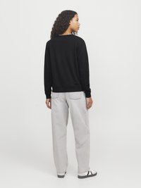 Een persoon met krullend haar staat met de rug naar ons gekeerd, gekleed in een zwart sweatshirt, lichtgrijze jeans en witte sneakers met zwarte strepen.