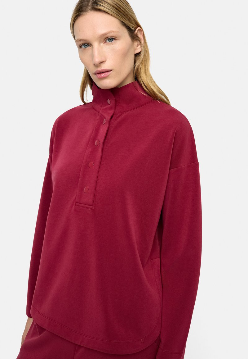 Kobieta o blond włosach w długim, bordowym swetrze z wysokim kołnierzem zapinanym na guziki oraz dopasowanych spodniach na białym tle.