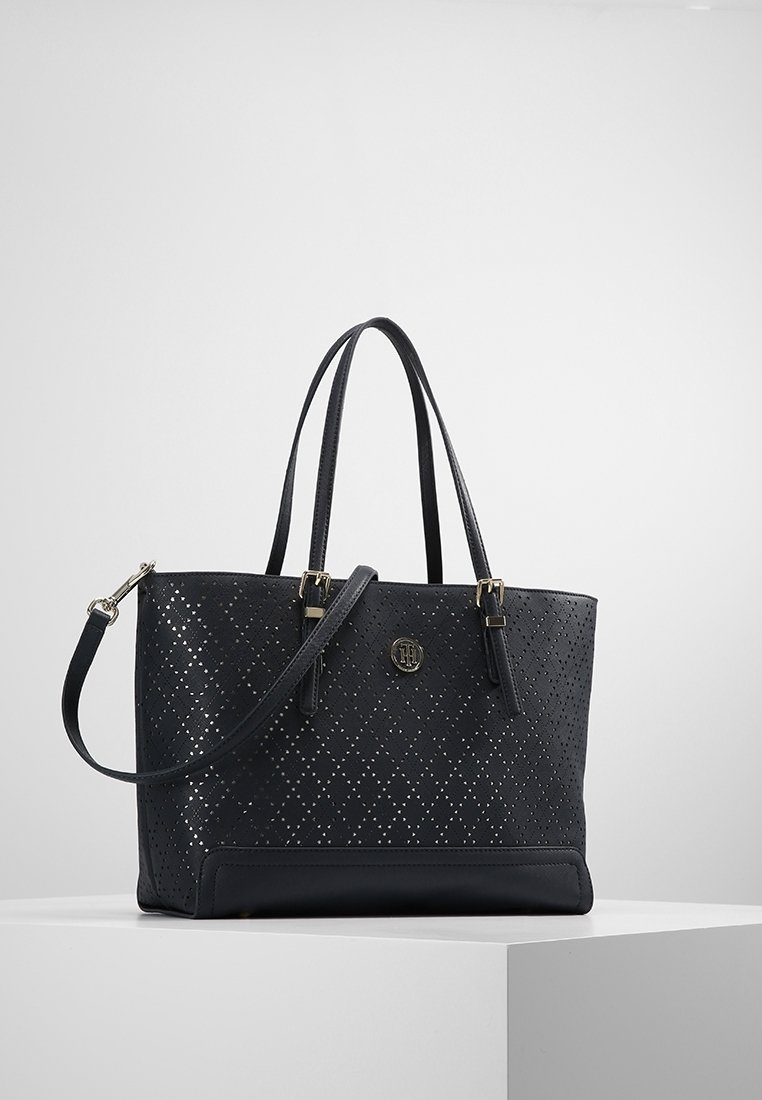 Sac cabas noir avec perforations en motif losange, deux bretelles d’épaule, une bandoulière amovible et un petit logo rond en métal sur le devant.