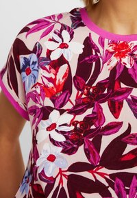 Närbild av en blommig mönstrad skjorta med vita, röda, lila och blå blommor samt matchande rosa kant på kragen och ärmarna.