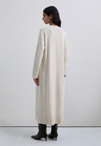 Vestido de punto color crema con una silueta larga, detalles acanalados en el dobladillo y hombros caídos. Llevado con botas negras hasta la rodilla.