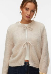 Jeune femme portant un cardigan en maille crème noué par deux nœuds sur un haut blanc et un jean bleu foncé, posant avec une expression neutre.