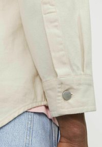 Cream-colored denim shirt featuring a folded cuff with a silver snap button, displayed over a light pink shirt and light blue denim.