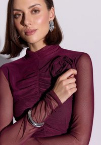 Bordowa dopasowana bluzka z marszczeniami i kwiatowym akcentem; przezroczyste rękawy; metalowa bransoleta na nadgarstek; gładka faktura i elegancki design.