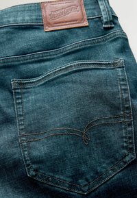 Jeanși din denim cu o spălare de culoare albastru închis, având o etichetă din piele maro și cusături de contrast pe buzunarul din spate.