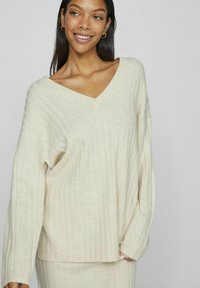 Suéter de punto acanalado color crema con escote en V y mangas anchas. Textura suave, corte relajado, combinado con una prenda inferior a juego.