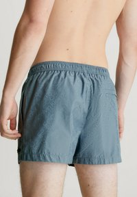 Pantaloni da bagno da uomo realizzati in tessuto grigio chiaro con un motivo logo su tutta la superficie, cintura elastica e tasca sul retro.