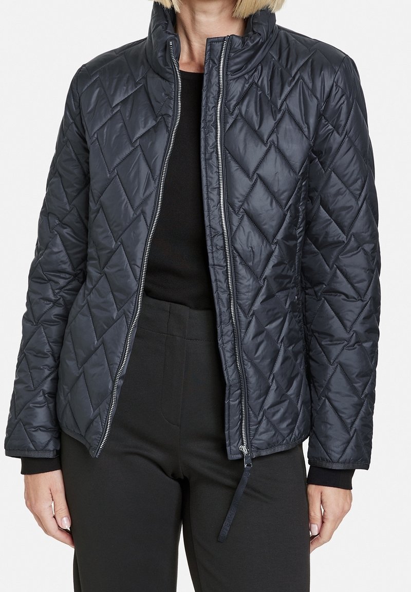 Femme portant une veste matelassée noire avec des coutures en losange, partiellement zippée, sur un haut noir et un pantalon noir.