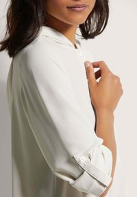 Witte knoopshirt gemaakt van gladde stof, met een kraag en opgerolde mouwen met knoopdetails. Close-up van de schouder en hand.