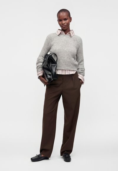 Personne debout, portant un pull gris sur une chemise à col, un pantalon marron, des chaussures noires, tenant un grand sac noir, posant avec assurance.