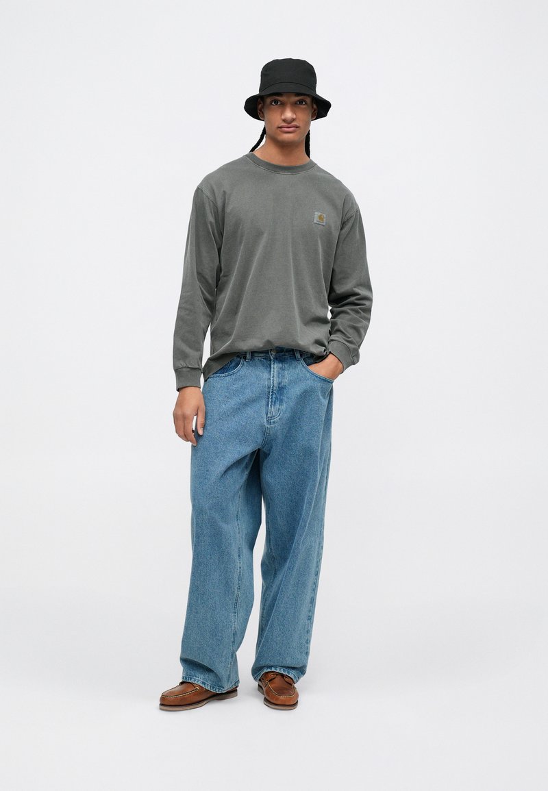 Grijze longsleeve shirt met logo, lichtblauwe baggy jeans, bruine instap schoenen en een zwarte bucket hat. Soepele texturen en een ontspannen pasvorm.