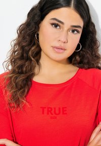Červené tričko s krátkým rukávem a kulatým výstřihem s textem "TRUE SIZE" v tmavším odstínu. Měkký, lehký materiál s volným střihem.