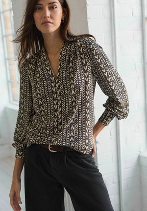 La blouse présente un motif marron et crème avec des motifs en chevrons et des feuilles, des manches longues, un col en V et un tissu léger. Portée avec un pantalon noir.