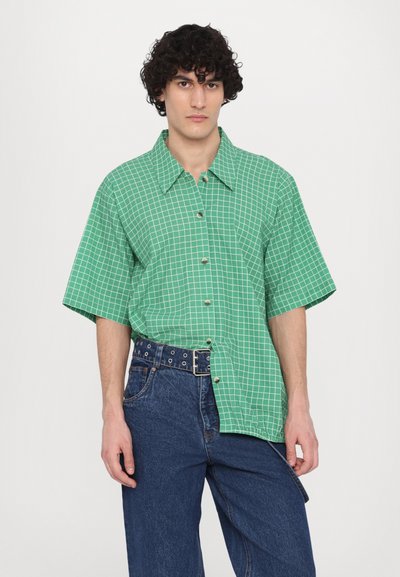 Green short-sleeve shirt with a grid pattern, featuring a collar and buttons. Paired with dark blue denim pants and a studded belt.