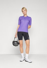 Kortermet lilla sykkeltrøye med glidelås, svarte sykkelshorts, hvite sokker og en svart hjelm holdt i hånden. Skoene er hvite med aksenter.