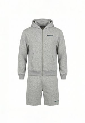 Mężczyzna zwrócony tyłem, ubrany w jasnoszarą bluzę z kapturem z napisem "closurelondon" i pasujące szorty, białe trampki na białym tle.