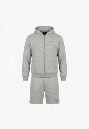 Mężczyzna zwrócony tyłem, ubrany w jasnoszarą bluzę z kapturem z napisem "closurelondon" i pasujące szorty, białe trampki na białym tle.