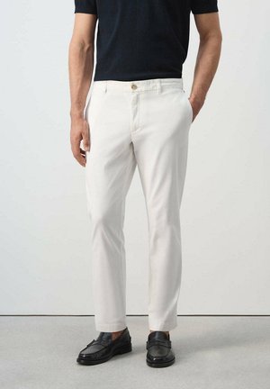 Hombre vestido con pantalones ajustados blancos, mocasines negros y una camisa negra de manga corta, de pie con una mano en el bolsillo.
