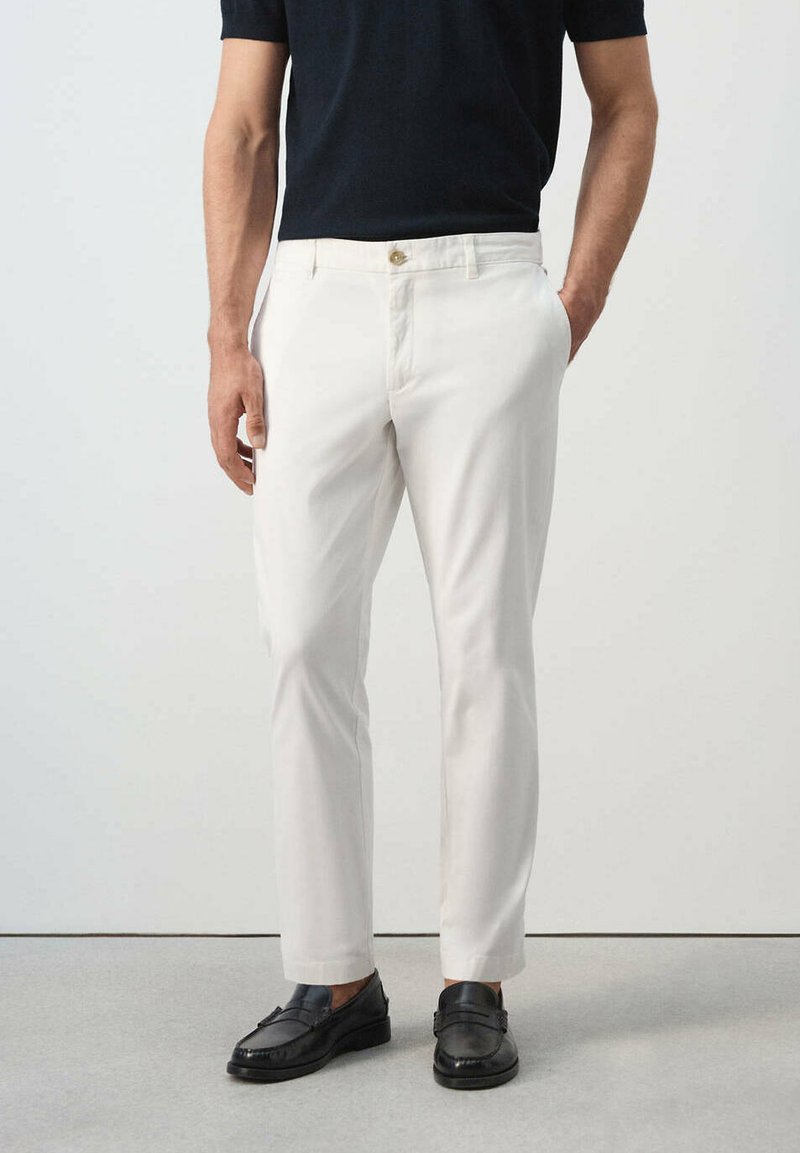 Homme portant un pantalon slim blanc, des mocassins noirs et une chemise noire à manches courtes, debout avec une main dans la poche.