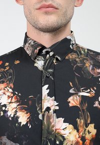 Bloemenoverhemd met knopen in zwarte stof met kleurrijke bloemenpatronen. Klassieke kraag en zwarte knopen. Soepele textuur, aansluitend ontwerp.