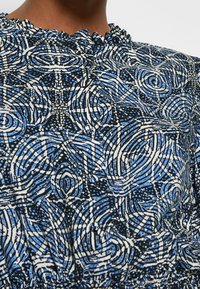 Blusa a fantasia in nero, blu e bianco. Presenta motivi vorticosi e una scollatura volant. Tessuto leggero con un aspetto texturizzato.
