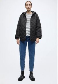 Μαύρο μπουφάν με καπιτονέ ραφή και κουκούλα, φορεμένο πάνω από ριγέ πουκάμισο. Το σύνολο ολοκληρώνεται με μπλε τζιν παντελόνι με ίσια γραμμή και μαύρα μποτάκια αστραγάλου.