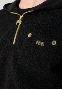 Pullover de fleece preto com fecho de zip, apresentando detalhes em dourado e um bolso no peito. Tecido macio e texturizado com capuz forrado.