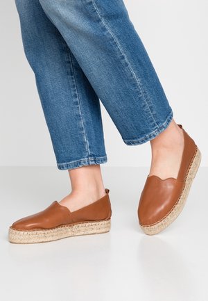 Persoon draagt cropped blauwe jeans en bruine leren slip-on espadrilles met gevlochten jute plateauzolen op een witte vloer.
