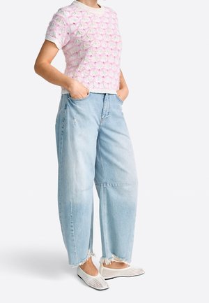 Person trägt ein pinkes kurzärmeliges Stricktop, weite hellblaue zerrissene Jeans und weiße Mesh-Slipper und steht mit den Händen in den Taschen.