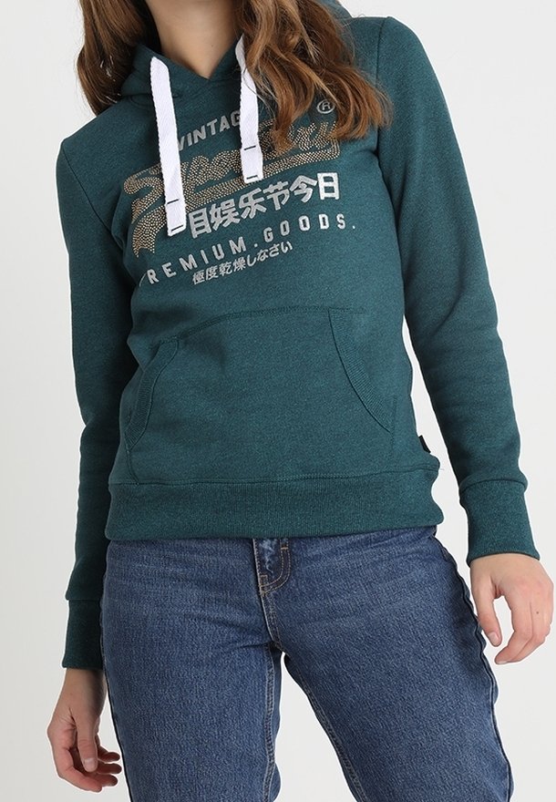 Felpa teal con lacci bianchi e tasca frontale. Caratterizzata da stampa di testo in oro e bianco. Abbinata a jeans blu, mostrata in un ambiente da studio.