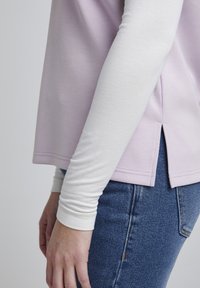 Top de manga corta en lavanda, superpuesto sobre una camiseta de manga larga blanca, con una abertura lateral y combinado con jeans de mezclilla azules.