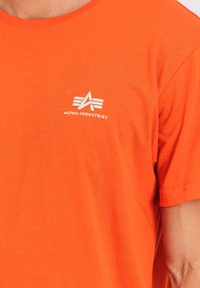 Kurzarm-T-Shirt aus leuchtend orangefarbenem Baumwollstoff mit rundem Halsausschnitt. Verfügt über ein kleines weißes Logo auf der linken Brustseite.