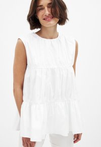 Valge ilma varrukateta pluus ümmarguse dekoltee, volangide ja laia lõikega, valmistatud siledast kangast, millel on veidi läikiv tekstuur.