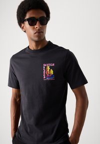 Sort bomulds T-shirt med et farverigt grafisk tryk af træer og teksten "SCOTCH AMSTERDAM" på venstre brystområde. Korte ærmer.