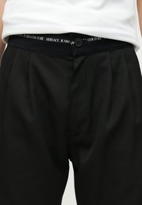 Κοντινή λήψη μαύρου παντελονιού με πιέτες και κλείσιμο με κουμπί, φορεμένο με λευκό τοπ που είναι μπουσουρεμένο μέσα, δείχνοντας τη ζώνη του Versace Jeans Couture.