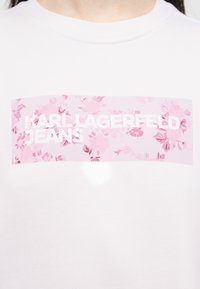 Λευκό πουκάμισο με ρόζ ορθογώνιο λουλουδάτο μπαλώμα που εμφανίζει το κείμενο "KARL LAGERFELD JEANS" σε λευκά κεφαλαία γράμματα στο στήθος.