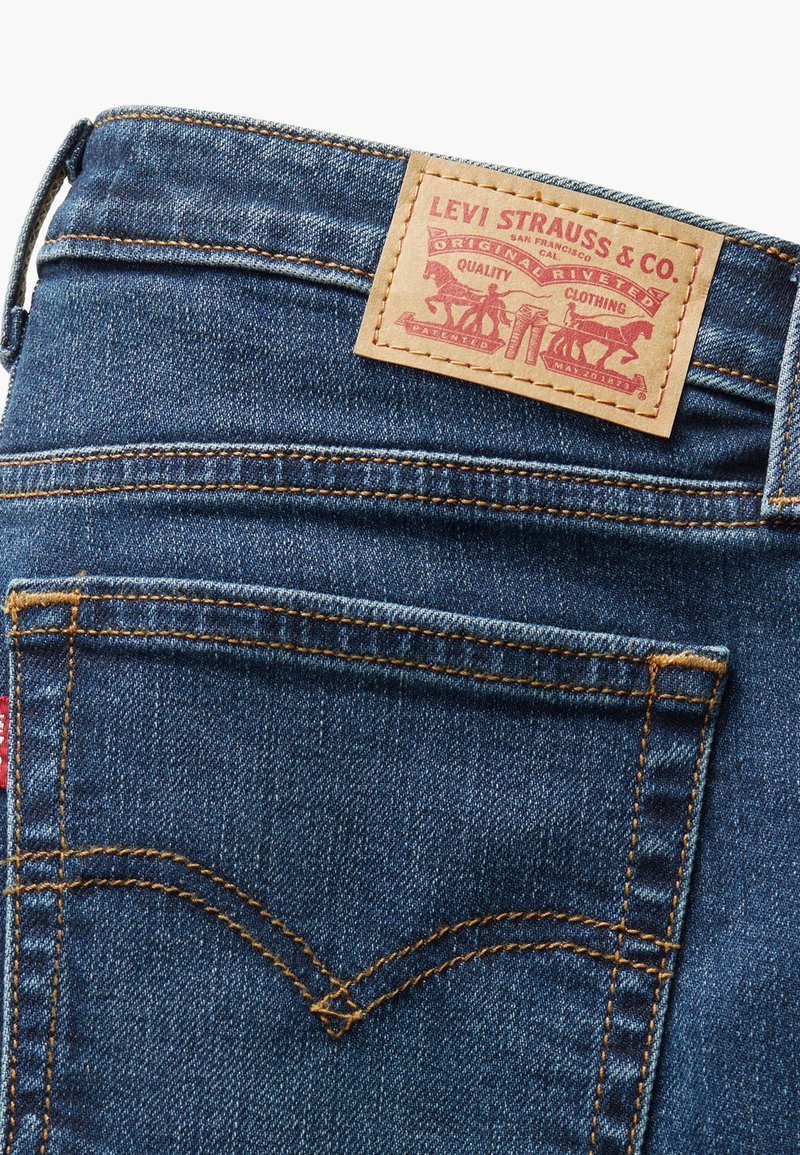 Jeans in denim blu scuro con una patch in pelle che presenta testo e grafiche rosse. Cuciture gialle e un distintivo motivo a V sulle tasche posteriori.