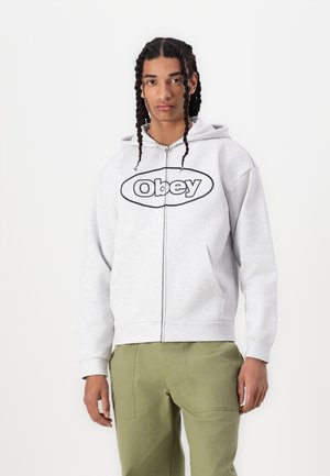 Felpa grigia con zip e logo "Obey" in nero, dotata di tasca frontale e cappuccio con cordino. Indossata con pantaloni verde chiaro.
