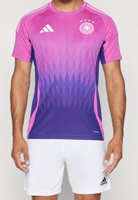 Mężczyzna w różowo-fioletowej koszulce piłkarskiej Niemiec z białym logo Adidas oraz białych szortach z logo Adidas.