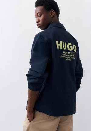 Hombre con chaqueta azul marino de mangas remangadas y logo "HUGO" impreso en la espalda, combinada con pantalones beige sobre un fondo liso.