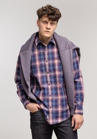 Koszula w kratę w kolorze granatowym, czerwonym i białym, z długimi rękawami i zapięta na guziki. Na ramionach leży jasnofioletowy sweter. Widać ciemne dżinsy.