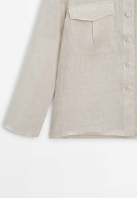 Chemise en lin beige à manches longues avec boutons frontaux et une seule poche poitrine avec rabat sur fond blanc.