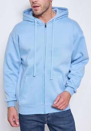 Homme portant un sweat à capuche zippé bleu clair avec poches avant et cordon de serrage à la capuche, associé à un jean bleu foncé.