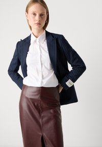 Nainen, jolla on päällään tummansininen pinstripe-puku, valkoinen napitettu paita ja korkea vyötärö burgundinpunainen nahkahame, jossa on etureikä, seisoo kädet lanteilla.