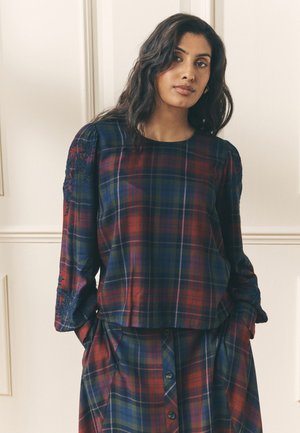 Woman with long dark hair wears a red, blue, and green plaid top and matching skirt, standing against a light-colored paneled wall.
