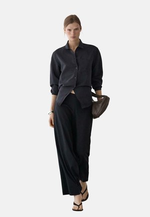 Kobieta ubrana w luźną czarną koszulę na guziki i szerokie spodnie, niosąca brązową torebkę, idzie do przodu z neutralnym wyrazem twarzy.